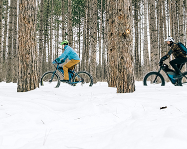 two people riding their bikes through a snowy forest